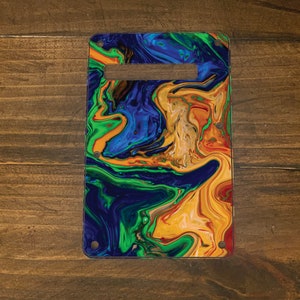 May include: Rectangular cutting board with a handle cutout, featuring a vibrant abstract design. The pattern blends shades of blue, green, orange, and yellow, creating a marbled effect. The board is secured with small silver rivets.