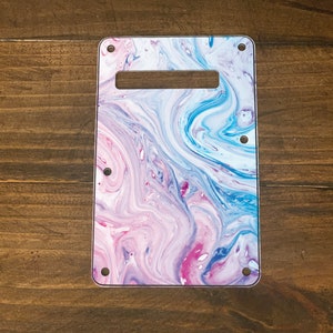 May include: A white guitar pickguard with a blue and pink marbled design. The pickguard has four screw holes.