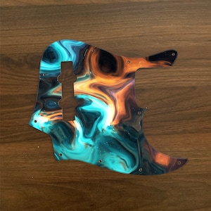 Puede incluir: Un golpeador de bajo con un patrón arremolinado de color turquesa, naranja y negro. El golpeador tiene un recorte para las pastillas y los mandos, y varios agujeros para tornillos. El diseño se asemeja a una nebulosa o un cielo en llamas.
