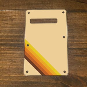May include: A cream-colored guitar pickguard with a diagonal stripe pattern in shades of brown, orange, and yellow.