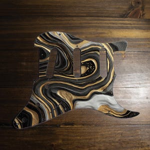 May include: A guitar pickguard with an abstract design. The pickguard has a swirling pattern of black, gold, and white, creating a marbled effect. It has cutouts for pickups and mounting screws, set against a wooden background.