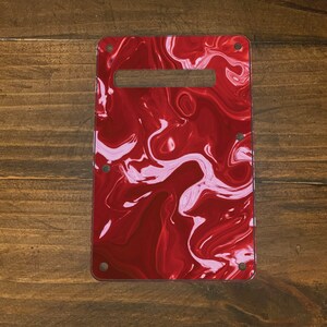 May include: Red and white swirl guitar pickguard with a rectangular cutout for a pickup. The pickguard has a glossy finish and is made of plastic.