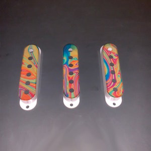 Puede incluir: Tres tapas de pastilla de guitarra con un patrón de remolino colorido en tonos de amarillo, naranja, rojo, azul, verde y morado. Las tapas son blancas con ribetes negros y tienen seis agujeros.