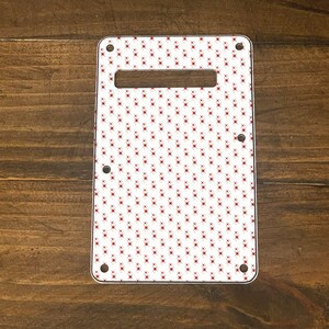 May include: White guitar pickguard with a red repeating pattern of small stars. The pickguard has four screw holes.