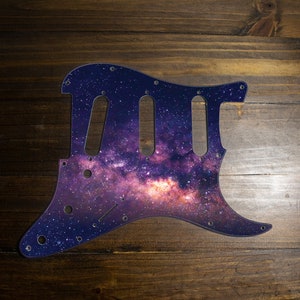 Puede incluir: Un golpeador de guitarra con un diseño de galaxia vibrante. El golpeador, diseñado para una guitarra eléctrica, presenta un fondo azul oscuro salpicado de estrellas blancas y una nebulosa central de tonos morados y rosas. Tiene agujeros pre-perforados para el montaje.
