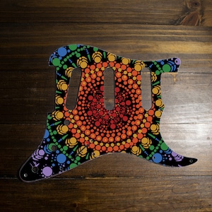 May include: A black guitar pickguard with a colorful mandala design. The mandala features a rainbow of colors, including red, orange, yellow, green, blue, and purple. The design is made up of small dots that create a swirling pattern.