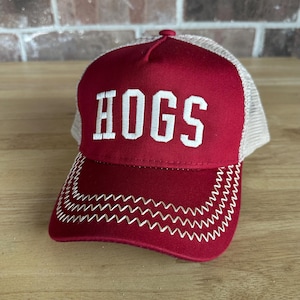 May include: A maroon trucker hat with the word "HOGS" embroidered in white block letters. The hat has a mesh back and a curved brim with white zigzag stitching.