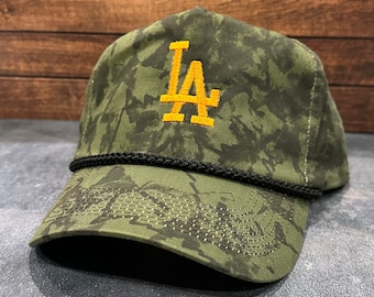 May include: A green camouflage baseball cap with a black rope trim and a yellow embroidered "LA" logo.