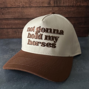May include: A baseball cap with a brown peak and a cream-coloured crown. The phrase "not gonna hold my horses" is embroidered in brown on the front. The cap has a curved peak and a button on top.
