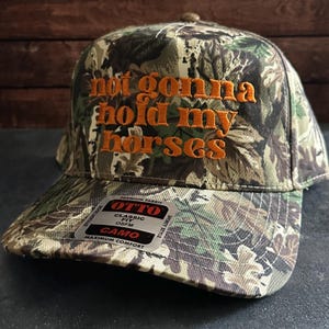May include: A camouflage baseball cap with the embroidered phrase "not gonna hold my horses" in orange thread. The cap has a curved peak and a tag with the brand name "OTTO" and the word "CAMO".