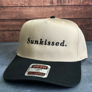 Op de afbeelding: Een beige baseballpet met een zwarte klep. De pet heeft zwarte borduurwerk met de tekst "Sunkissed." De pet heeft een label aan de zijkant met de tekst "Otto" en "Classic Fit OS FM" en "Mid Profile Maximum Comfort."