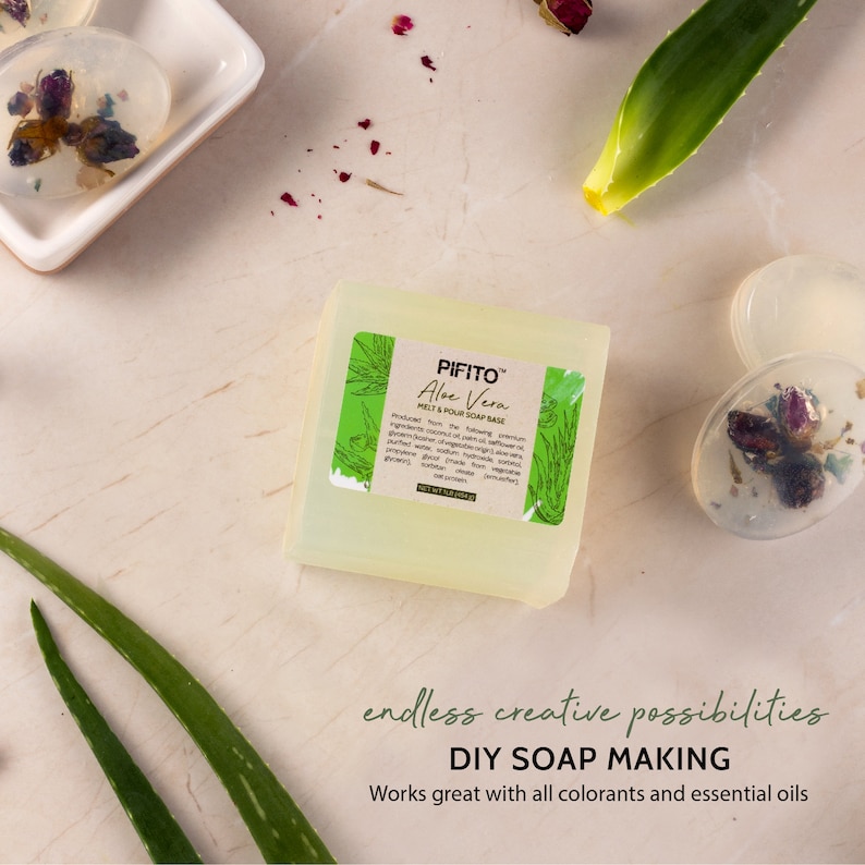May include: A clear, rectangular bar of soap base with a green label that reads "PIFITO&trade; Aloe Vera MELT & POUR SOAP BASE". The soap base is sitting on a light brown surface with green aloe vera leaves and red flower petals scattered around it.