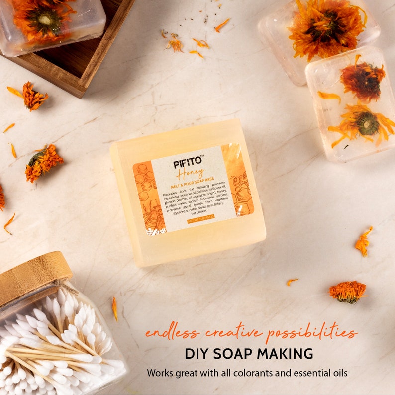 Op de afbeelding: Een transparante, rechthoekige blok van smelt- en gietzeepbasis met een label met de tekst "PIFITO Honey Melt & Pour Soap Base". De zeepbasis ligt op een wit oppervlak met verspreide gedroogde bloemen. De tekst "endless creative possibilities DIY SOAP MAKING Works great with all colorants and essential oils" staat onder de zeepbasis.