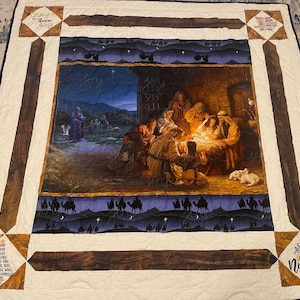 May include: A quilted blanket featuring a Nativity scene with a warm, inviting glow. The central image depicts the birth of Jesus, surrounded by figures in robes. The quilt has a cream-colored background with brown wood-like accents and decorative corner squares with text.