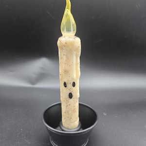 May include: A cream-colored, battery-operated candle with a yellow flame-shaped tip. The candle has a ghost face design and sits in a black metal holder. The candle is approximately 6 inches tall.