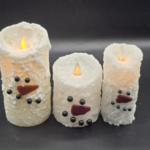 May include: Three white snowman-shaped candles with black eyes, red noses, and black buttons. The candles are lit and have a flickering flame.