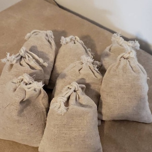 May include: Eight small, beige, drawstring fabric bags made of a natural material. The bags are arranged in a semi-circle on a beige surface.