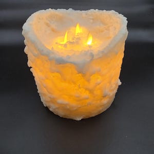 May include: A white pillar candle with a textured surface and three flickering flames. The candle is lit and casting a warm glow.