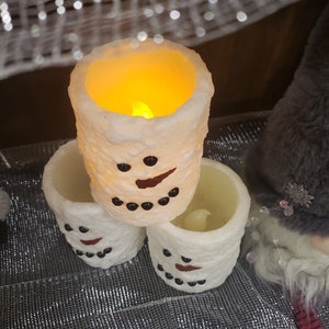 May include: Three white snowman-themed LED candles. Each candle has a textured surface, black dot eyes, and a red curved mouth. The top candle glows with a warm yellow light, creating a festive holiday ambiance.
