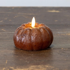 May include: A brown, pumpkin-shaped candle with a flickering flame. The candle has a textured surface and is set against a dark wooden background. The candle's warm glow suggests a cozy, autumnal theme.
