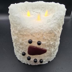 May include: A white snowman candle with a red nose and black eyes. The candle has three flickering LED lights.