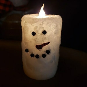 May include: A white snowman-shaped candle with a flickering LED flame. The candle features black dot eyes and a coal-like mouth and buttons. The candle is designed to resemble a snowman and is suitable for holiday decor.