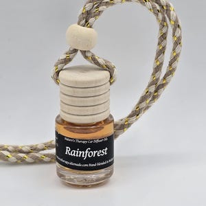 May include: A brown glass bottle with a black label that reads "Nature's Therapy Car Diffuser Oils Rainforest aromatherapy.idlemade.com Hand-Blended in Waikoloa" hanging from a beige and gold cord.