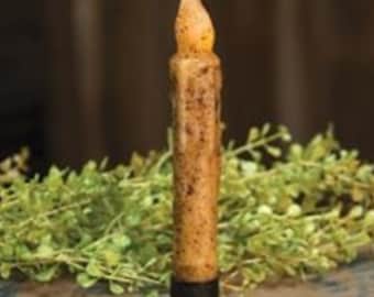 May include: A tall, battery-operated candle with a flickering amber flame. The candle has a textured, brown wax appearance with dark speckles and sits on a black metal base. Green foliage is in the background, creating a rustic aesthetic.