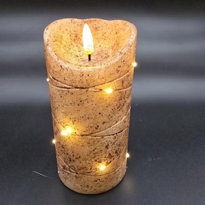 May include: A cylindrical, battery-operated candle with a textured, speckled surface. The candle has a flickering LED flame and is wrapped with a string of small, warm white lights. The candle is approximately 6 inches tall.