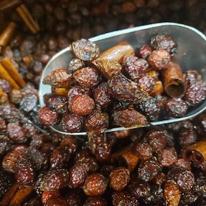 May include: A close-up of a scoop filled with dried rose hips and cinnamon sticks. The rose hips are dark brown and shiny, while the cinnamon sticks are light brown and have a rough texture.