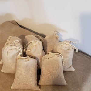 May include: Six small, off-white cotton drawstring bags filled with a beige material. The bags are arranged in a cluster on a brown burlap surface.