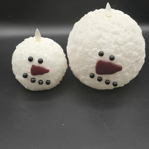 May include: Two white snowman-shaped candles with black eyes, red noses, and black buttons. The larger snowman is on the right and the smaller snowman is on the left.