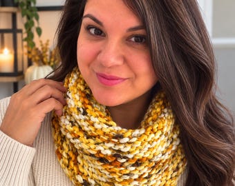 May include: A chunky knit infinity scarf featuring a variegated color palette of mustard yellow, cream, white, and dark brown yarn. The scarf is constructed with a thick, textured stitch pattern that creates a dense, layered appearance around the neck.