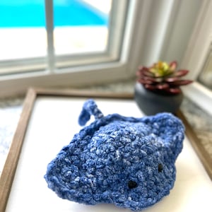 May include: A blue crocheted stingray with black eyes. The stingray is sitting on a white surface.