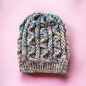 Könnte beinhalten: Eine bunte Strickmütze mit einem Zopfmuster. Die Mütze ist aus verschiedenen Blautönen, Rosa- und Gelbtönen gefertigt.