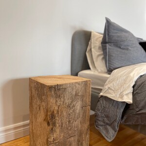 May include: A rustic wooden end table with a natural wood finish. The table is square and has a simple design. It is sitting on a hardwood floor next to a bed with grey bedding.