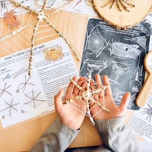 May include: A handmade spider web craft made with sticks and yarn, held in open hands. Instructions and diagrams for making spider webs are visible on the table, along with a wooden magnifying glass and spider illustrations.