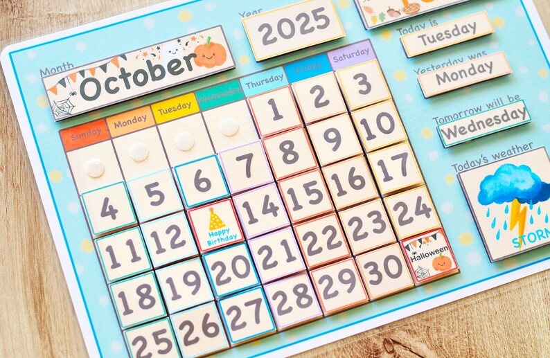 May include: A colorful calendar with a blue background and a white frame. The calendar is for October 2025 and features a variety of illustrations, including a pumpkin, a birthday hat, and a Halloween pumpkin. The calendar is made of felt and has a velcro backing.