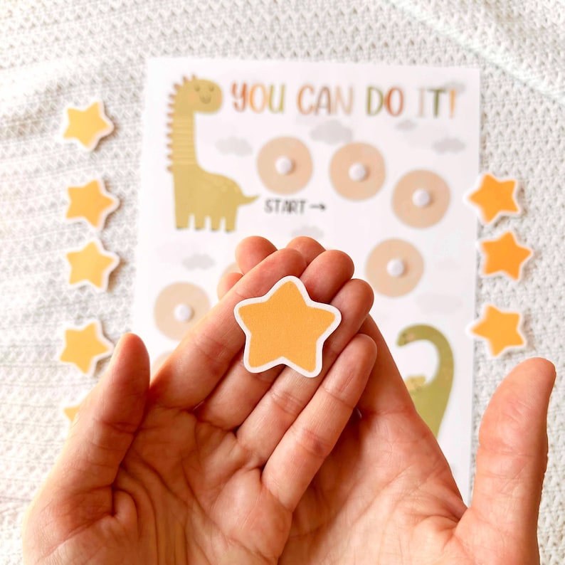 May include: A reward chart with a dinosaur illustration and the text "YOU CAN DO IT!". The chart has spaces for stickers, and several orange star stickers are scattered around. One star is held in a hand.