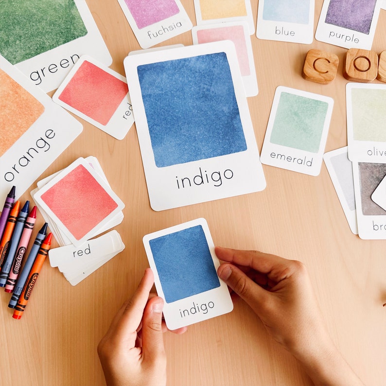 May include: A set of color cards with watercolor swatches and the color names written below. The colors include green, orange, red, fuchsia, blue, purple, emerald, olive, indigo, and brown. A hand is holding a card with a blue watercolor swatch and the word "indigo" written below.