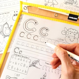 Peut inclure: Feuilles d'activit&eacute;s &eacute;ducatives pour tracer des lettres et dessiner. La photo montre une main tenant un marqueur blanc, tra&ccedil;ant la lettre "C" sur une feuille de travail. La feuille de travail contient les mots "cow" et "cat". Les feuilles sont dans un classeur en plastique transparent &agrave; bordure jaune.