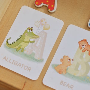 May include: Two flashcards with illustrations of an alligator and a bear. The alligator is green and holding balloons. The bear is brown and is sleeping on a large letter B. The words "ALLIGATOR" and "BEAR" are printed on the cards.