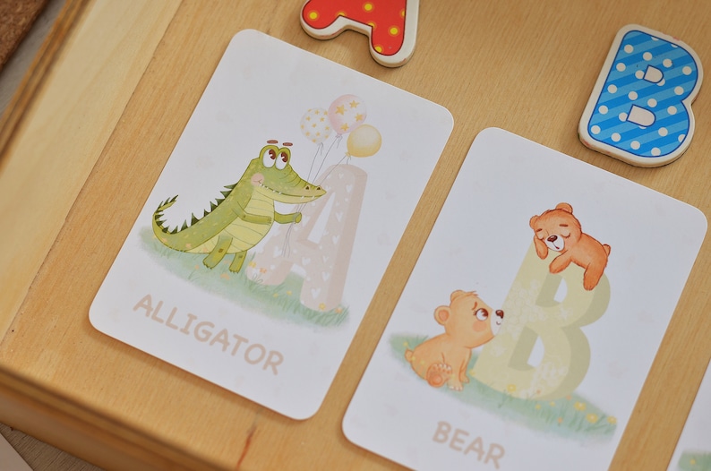 May include: Two flashcards with illustrations of an alligator and a bear. The alligator is green and holding balloons. The bear is brown and is sleeping on a large letter B. The words "ALLIGATOR" and "BEAR" are printed on the cards.