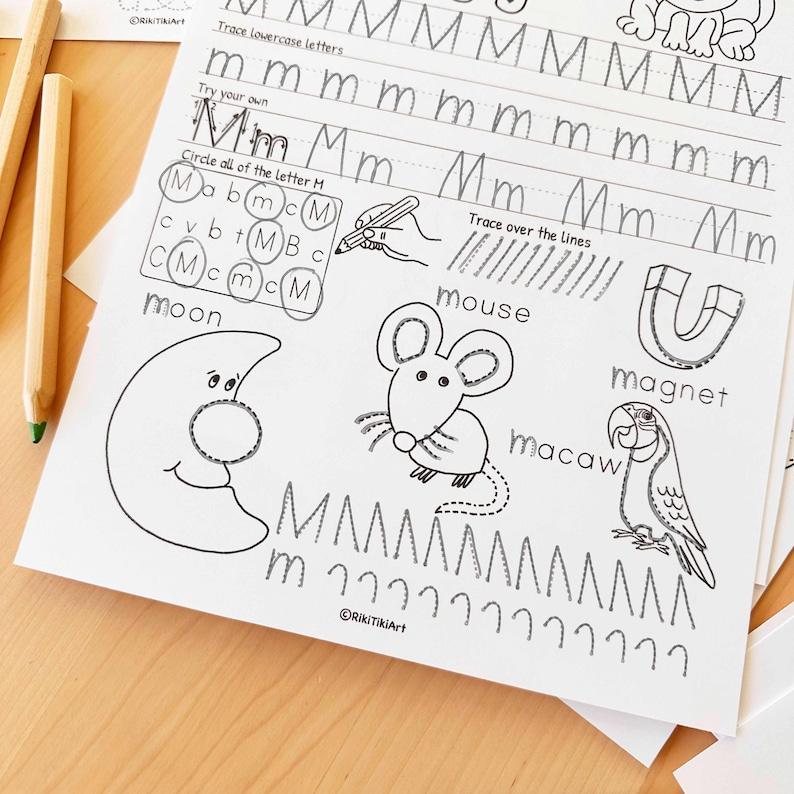 Peut inclure: Une feuille de travail blanche avec des exercices d'&eacute;criture pour la lettre "M". La feuille comprend des exercices de tra&ccedil;age, des illustrations d'une lune, d'une souris et d'un ara, et les mots "moon", "mouse", "magnet" et "macaw". Deux crayons sont visibles.