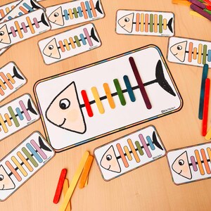 May include: A colourful fish-shaped card with a rainbow of lolly sticks glued to the outline of the fish's skeleton. The card is surrounded by other fish-shaped cards with lolly sticks glued to them.