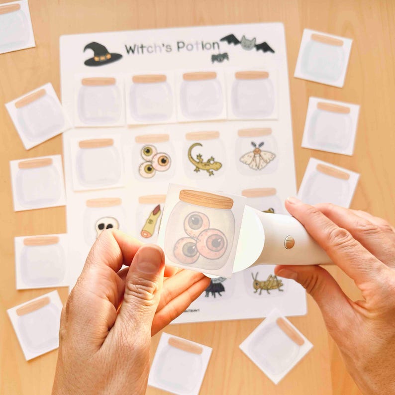 Puede incluir: Una hoja blanca titulada Witch's Potion con una cuadr&iacute;cula de frascos ilustrados, algunos vac&iacute;os y otros con elementos como globos oculares, un lagarto y una polilla. Una luz blanca de mano ilumina una tarjeta cuadrada con un frasco.