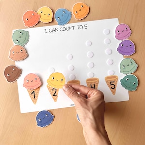 May include: A white educational board with the text "I CAN COUNT TO 5" and colorful ice cream cone-shaped number pieces. The numbers 1, 2, 4, and 5 are printed on cones. A hand is placing a yellow ice cream scoop on cone number 2.