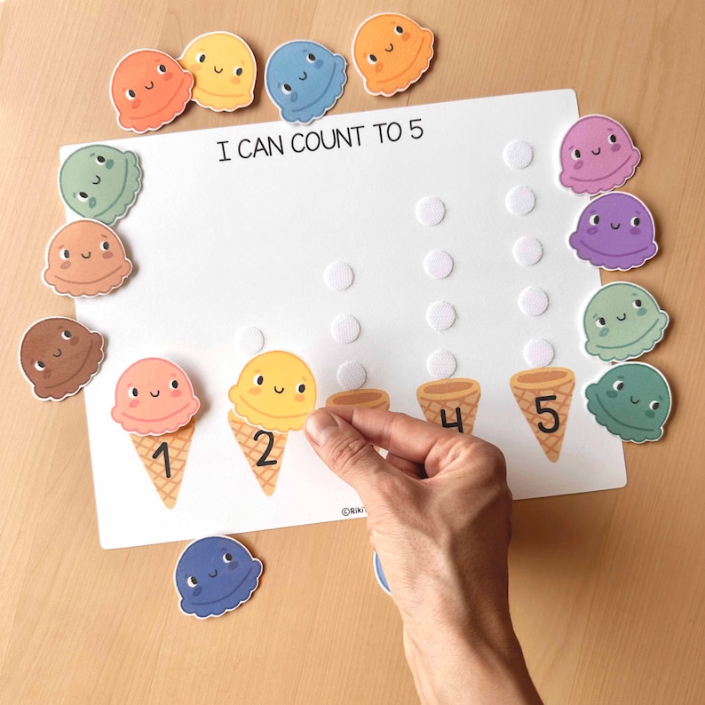 May include: A white educational board with the text "I CAN COUNT TO 5" and colorful ice cream cone-shaped number pieces. The numbers 1, 2, 4, and 5 are printed on cones. A hand is placing a yellow ice cream scoop on cone number 2.