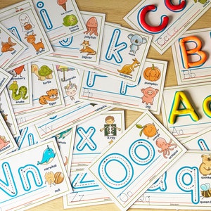May include: A set of alphabet flashcards with colorful illustrations and dotted lines for tracing letters. Each card features a different animal or object, such as a frog, a fish, a turtle, a tiger, a jellyfish, a kangaroo, a pumpkin, a pig, a duck, a dinosaur, a narwhal, a nut, a whale, an orange, an octopus, a quokka, a queen bee, a lemon, and a lion. The cards are printed on white paper with blue and red borders.