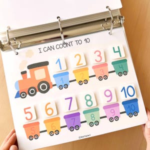 May include: A colorful educational counting activity. The page features a train with numbered train cars from 1 to 10. The text "I CAN COUNT TO 10" is at the top. The page is held open by a binder.